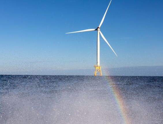 a faraway shot of a wind turbine in the ocean, at the bottom of the image there is mist coming of the water and a rainbow can be seen in the mist